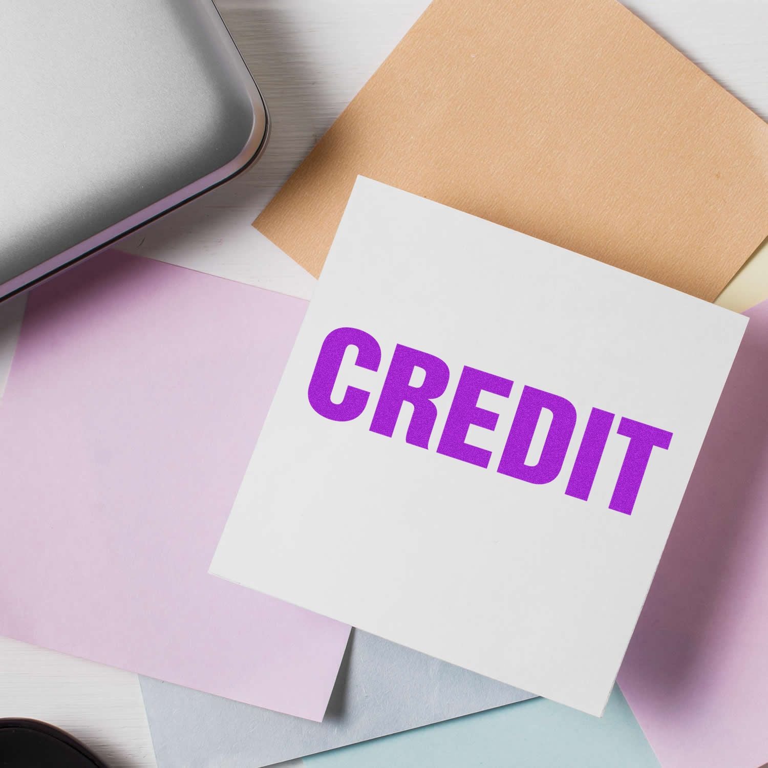 Bold red credit rubber stamp on a white paper surrounded by pastel-colored sheets and a silver laptop on a white desk.