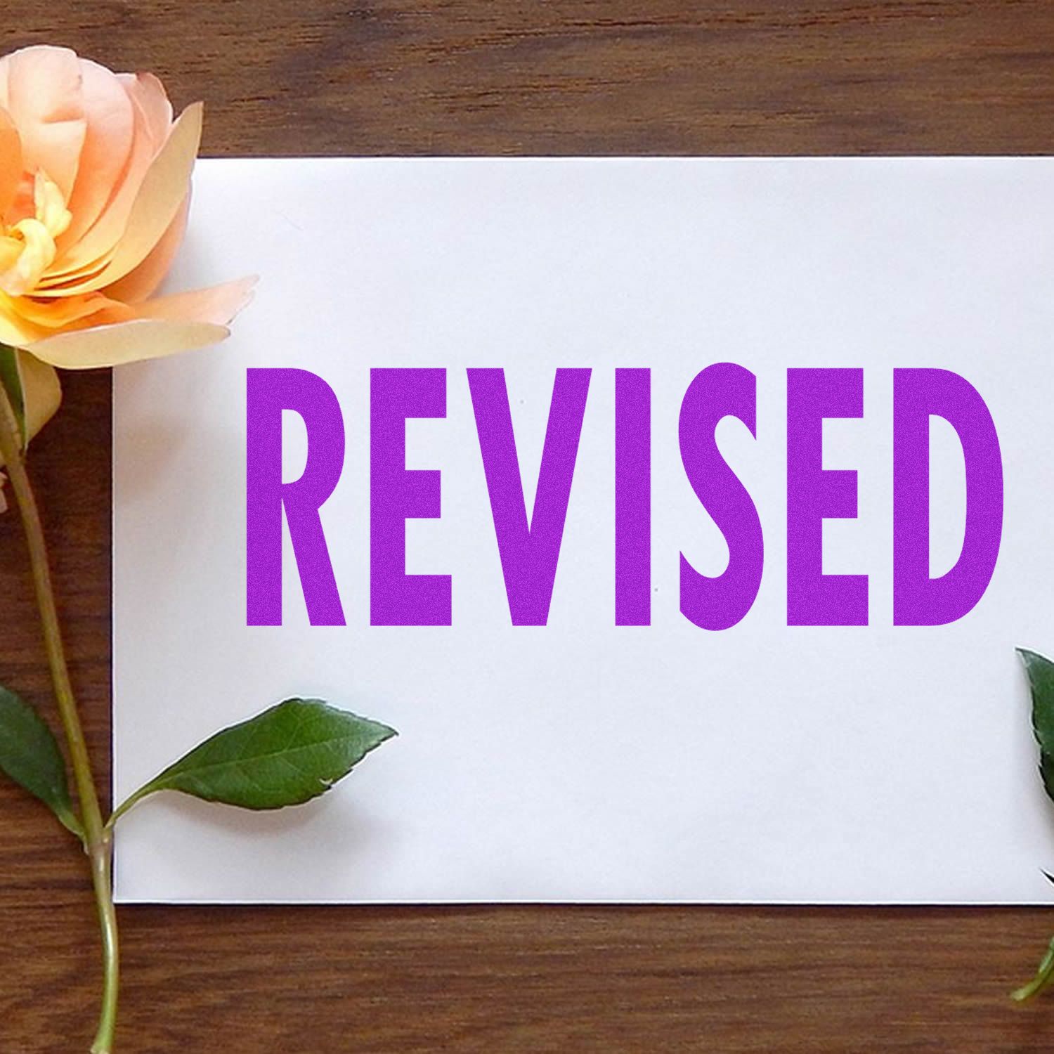 White paper stamped with a bold purple 'REVISED' rubber stamp, surrounded by a yellow rose and green leaves on a wooden surface.