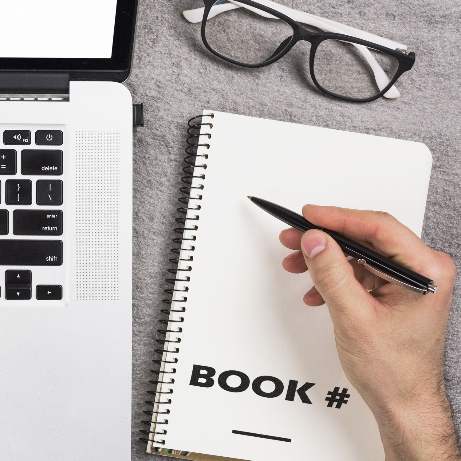 Hand holding a pen over a notebook stamped with BOOK # using the Book Rubber Stamp, next to a laptop and glasses on a gray surface.