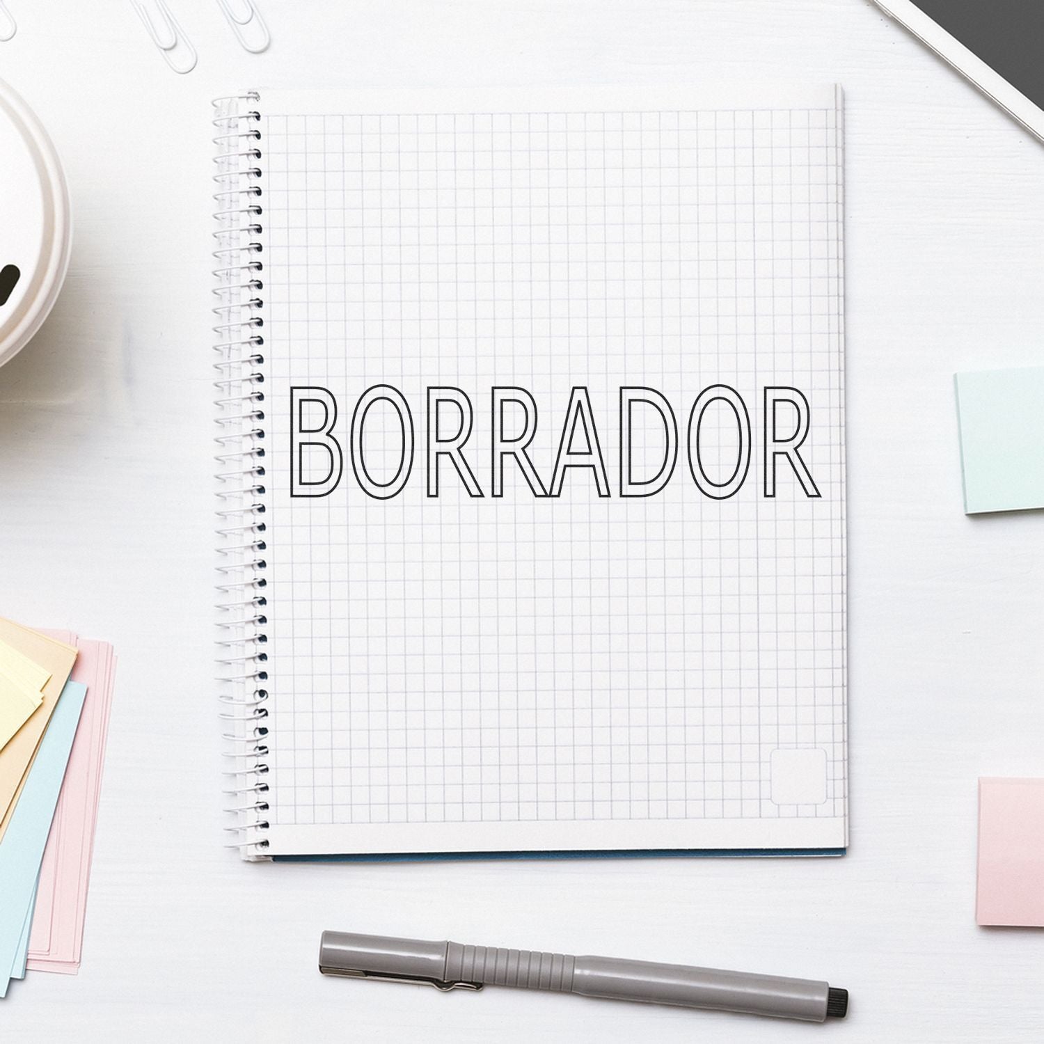 A notebook with grid paper stamped with BORRADOR using the Large Borrador Rubber Stamp, surrounded by a pen, sticky notes, and a tablet.