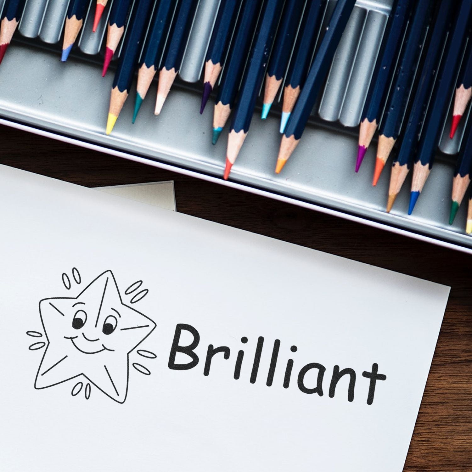 A piece of paper stamped with a smiling star and the word Brilliant using the Large Self Inking Brilliant Stamp, surrounded by colored pencils in a metal case on a wooden surface.