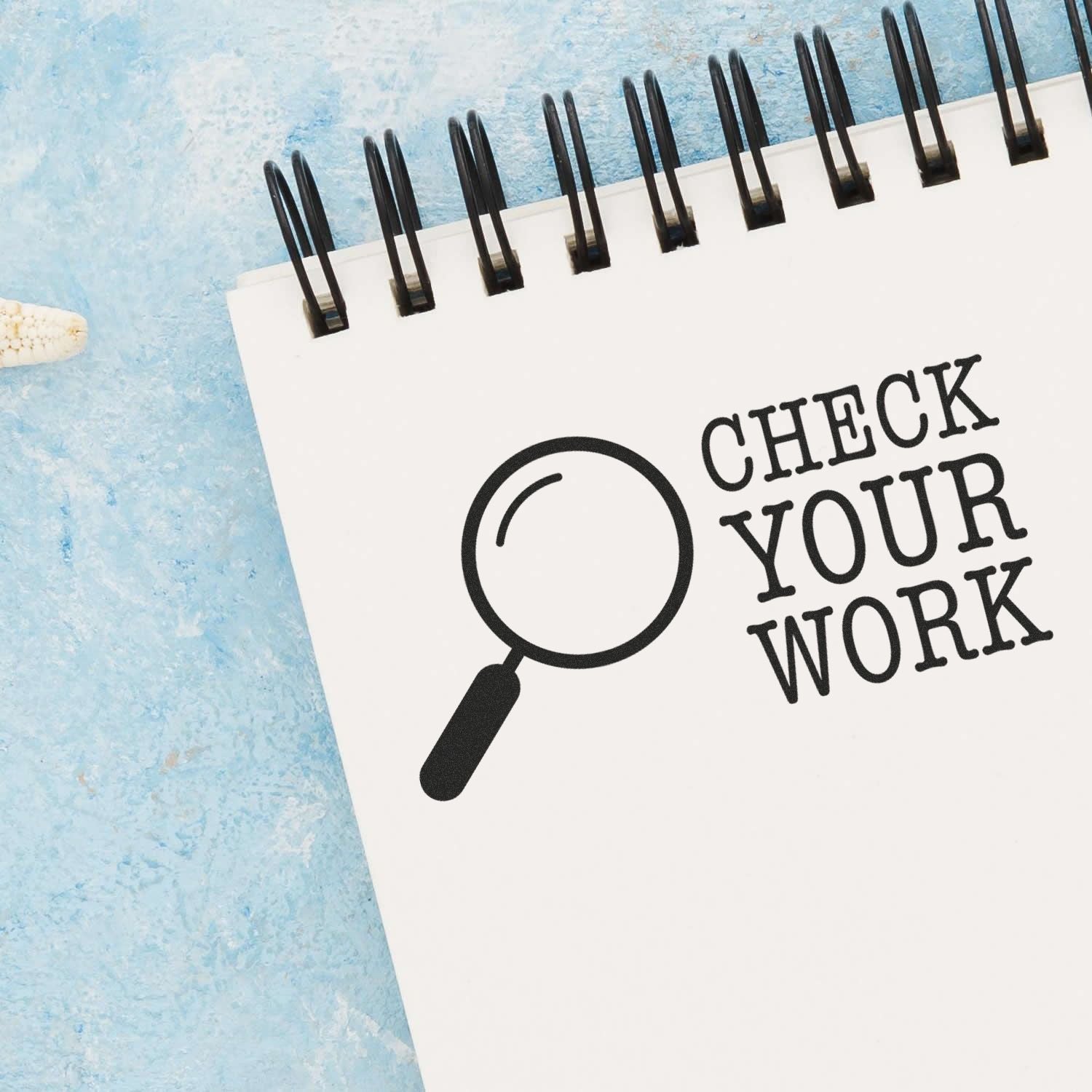 A notebook with a stamped message 'CHECK YOUR WORK' using the Large Self Inking Check Your Work Stamp, featuring a magnifying glass icon, placed on a light blue textured surface.