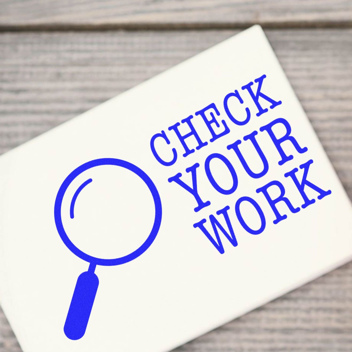 A close-up of the Large Self Inking Check Your Work Stamp, featuring a blue magnifying glass icon and the text CHECK YOUR WORK on a white background, placed on a wooden surface.