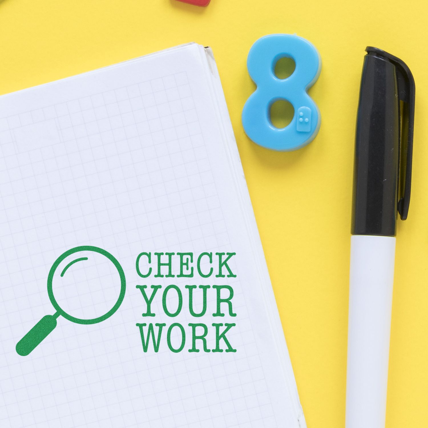 Large Self Inking Check Your Work Stamp in green ink on graph paper, next to a black pen and a blue number 8 eraser, all placed on a yellow background.