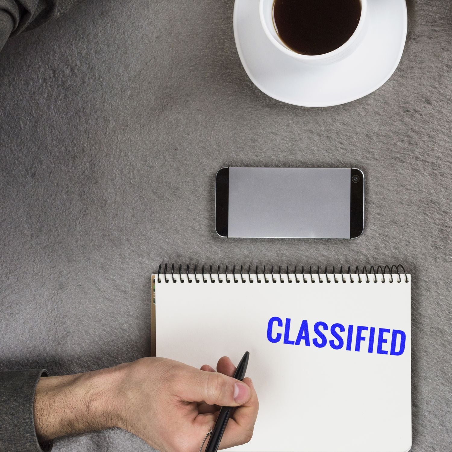 A hand writing in a notebook stamped with CLASSIFIED in blue ink using a Classified Rubber Stamp, with a phone and coffee cup nearby.