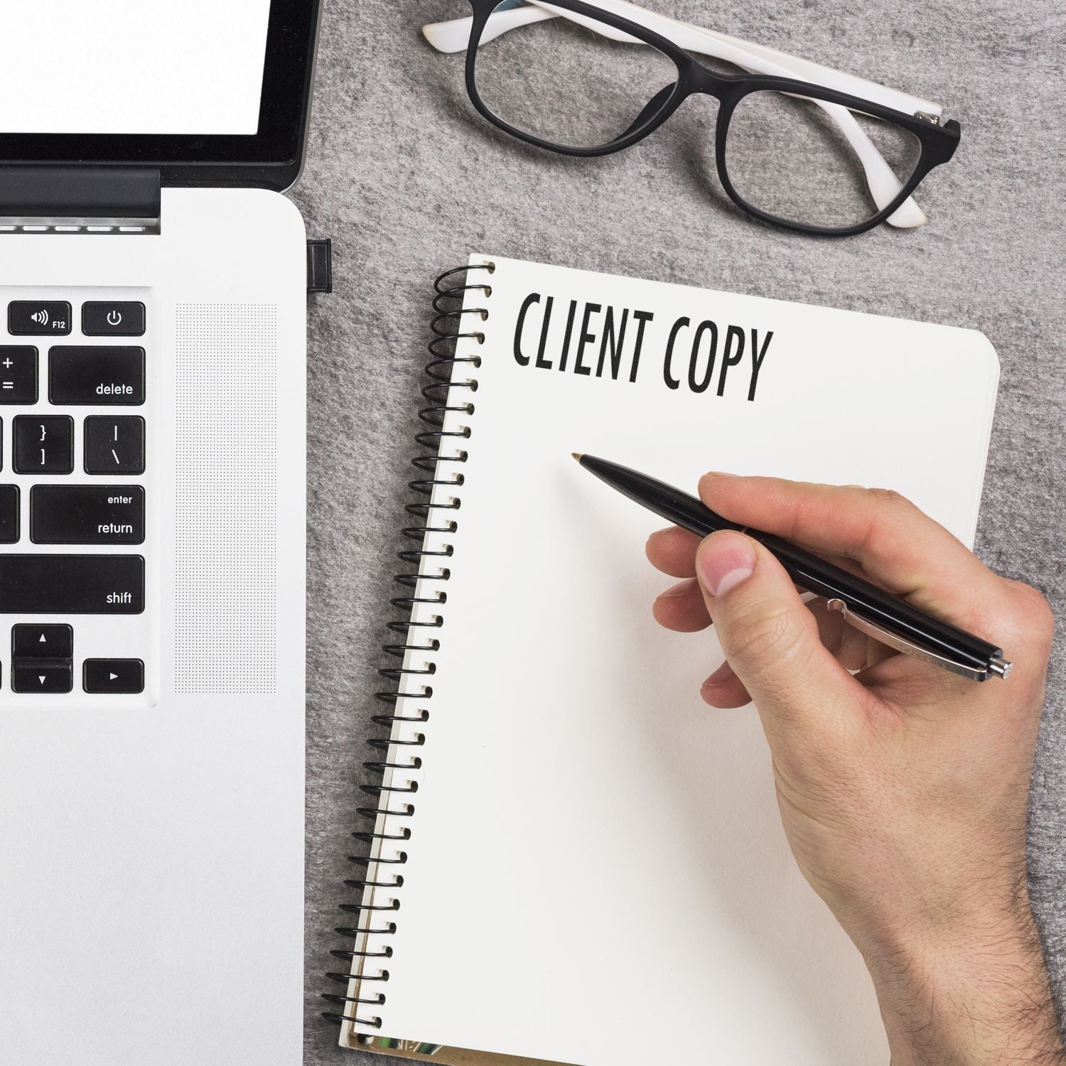 Hand holding a pen next to a laptop and glasses, writing CLIENT COPY on a notebook with a Client Copy Rubber Stamp nearby.