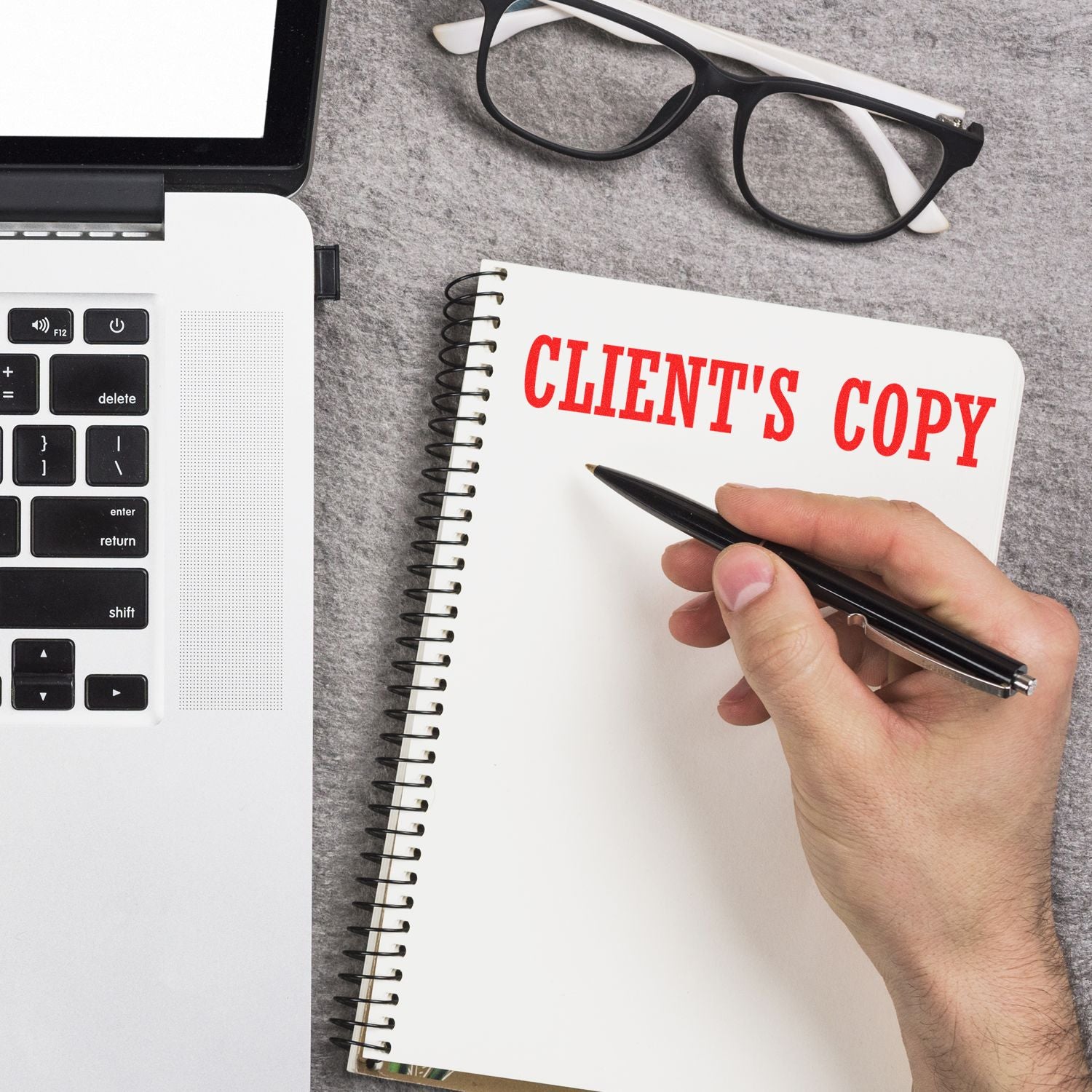 A hand holding a pen next to a laptop and glasses, with a notebook stamped CLIENT'S COPY using the Large Self Inking Clients Copy Stamp.