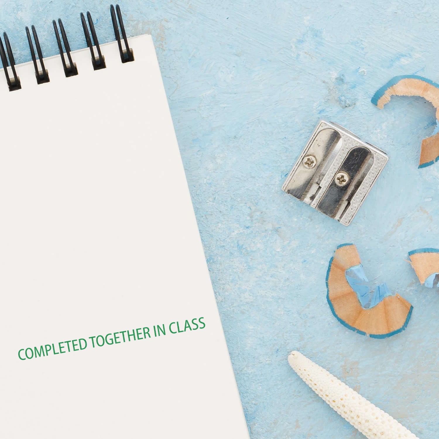 A notebook stamped with Completed Together In Class Rubber Stamp in green ink, next to a sharpener and pencil shavings on a blue surface.