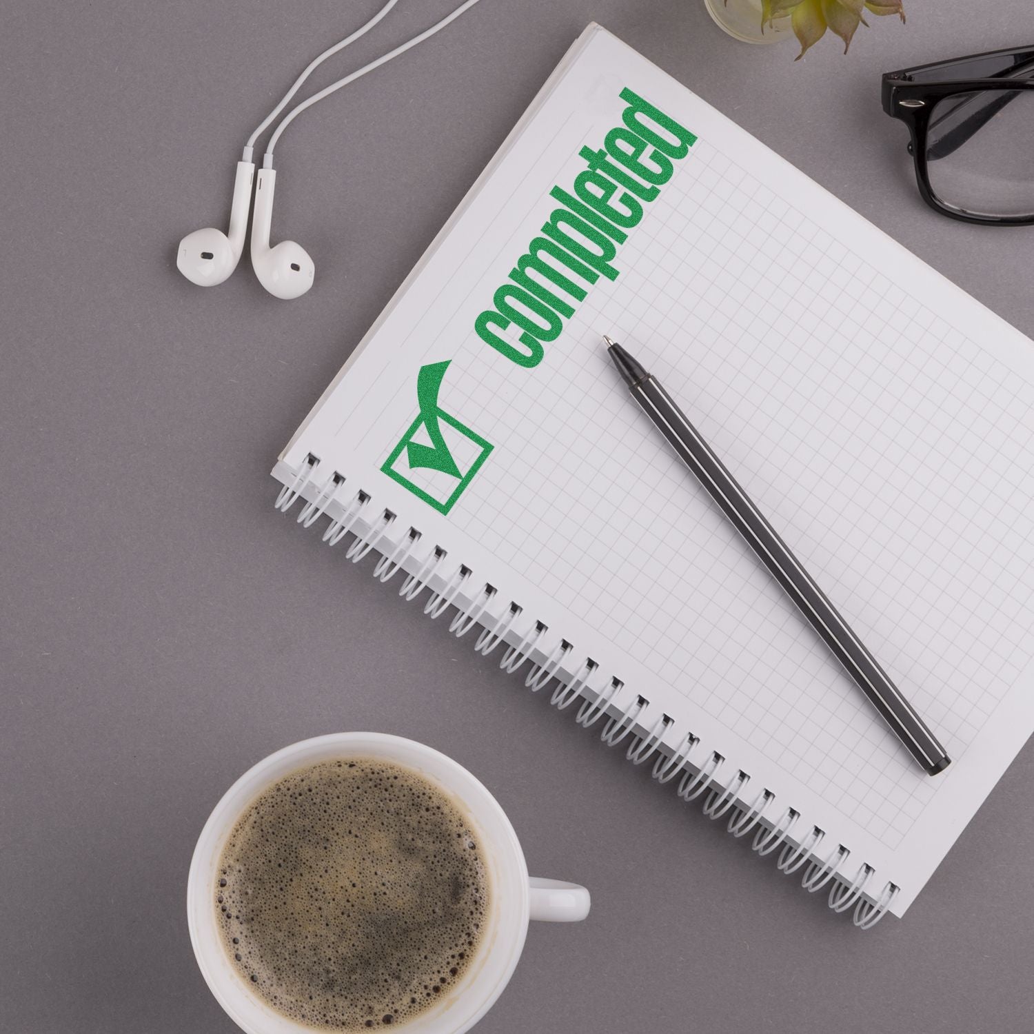 A notebook stamped with completed using the Large Completed with Checkbox Rubber Stamp, surrounded by a pen, coffee cup, glasses, and earphones.