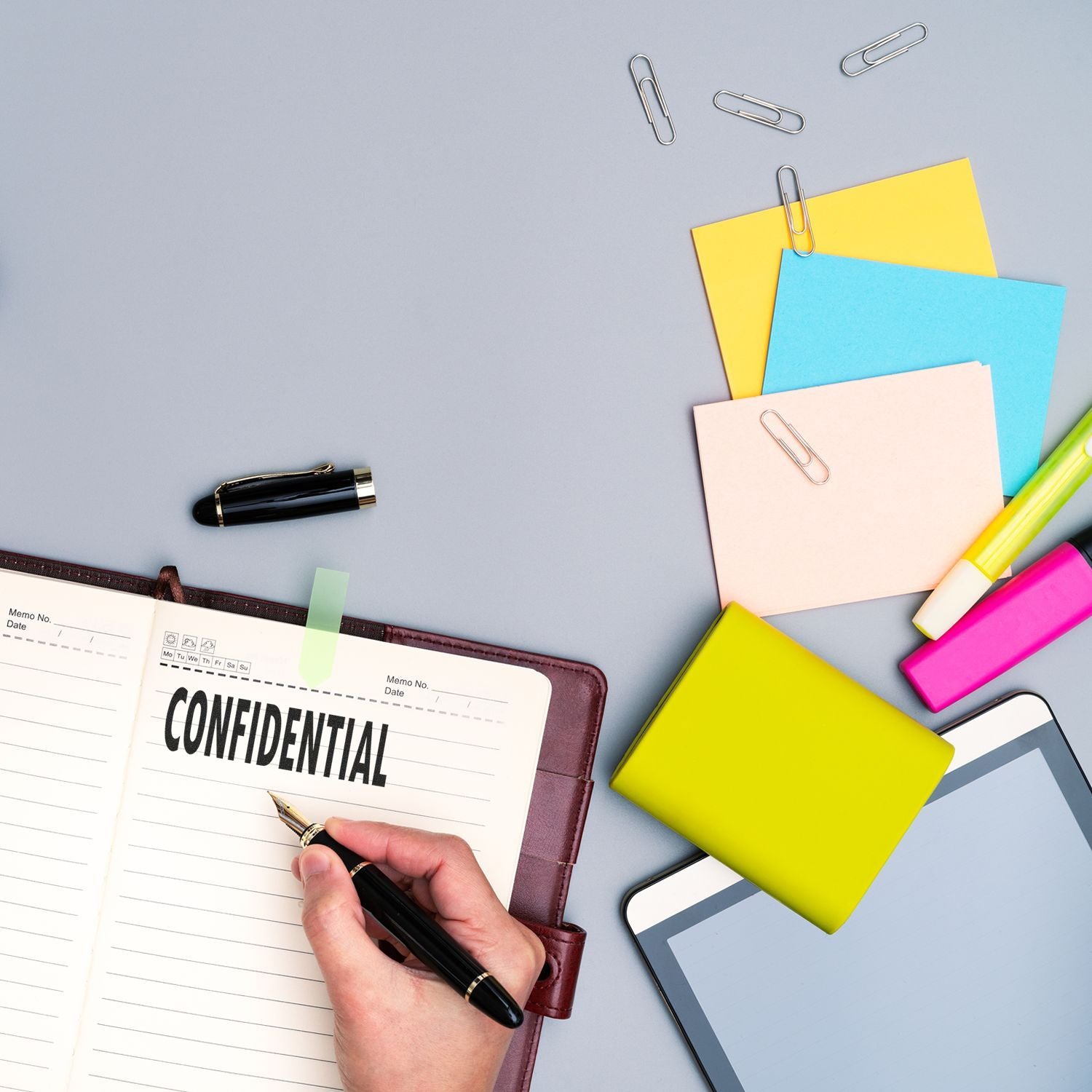 Hand using Large Confidential Rubber Stamp on a notebook, surrounded by colorful sticky notes, paper clips, pens, and a tablet.