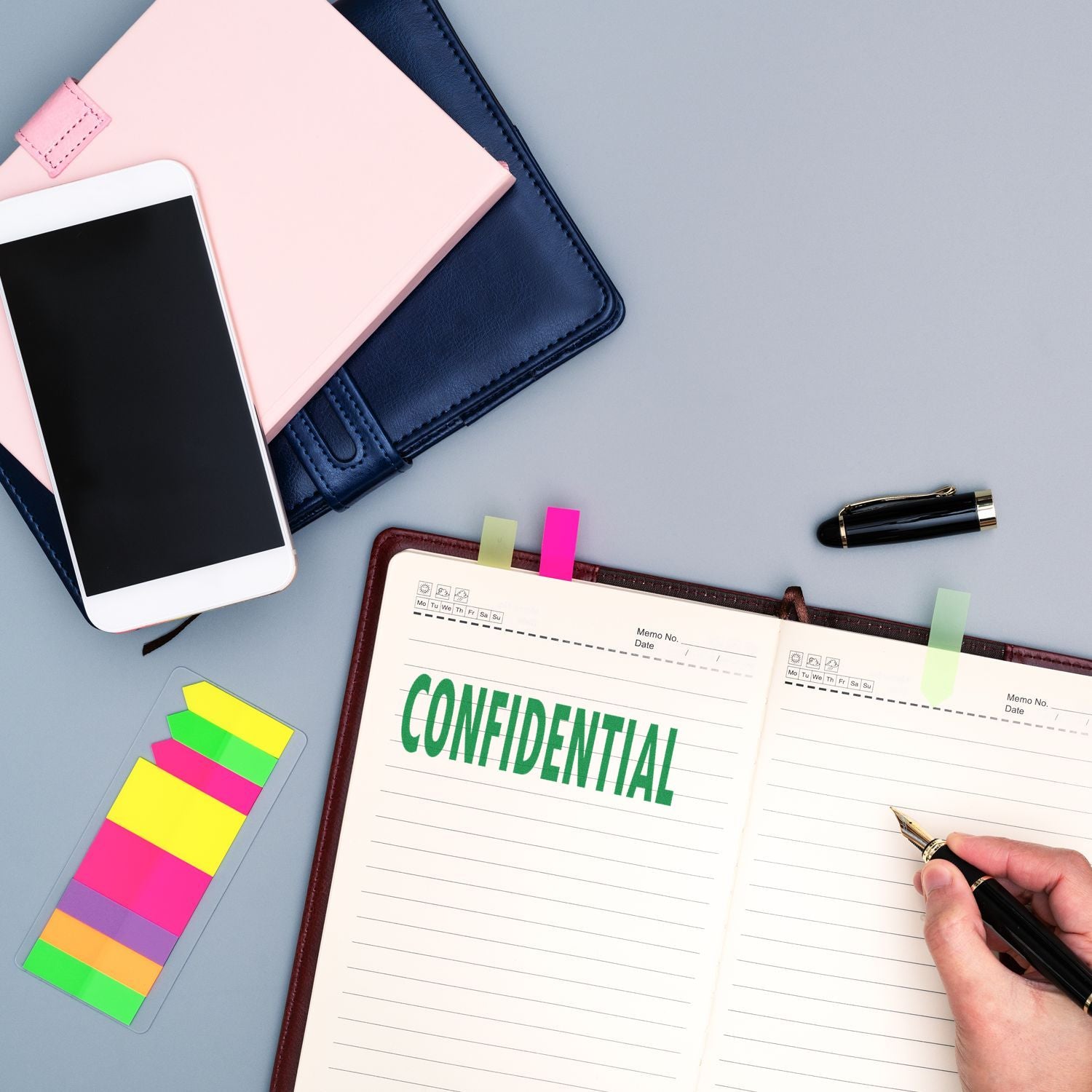 A desk with a notebook stamped CONFIDENTIAL using the Large Self Inking Confidential Stamp, surrounded by a smartphone, colorful sticky notes, a pen, and two closed notebooks.