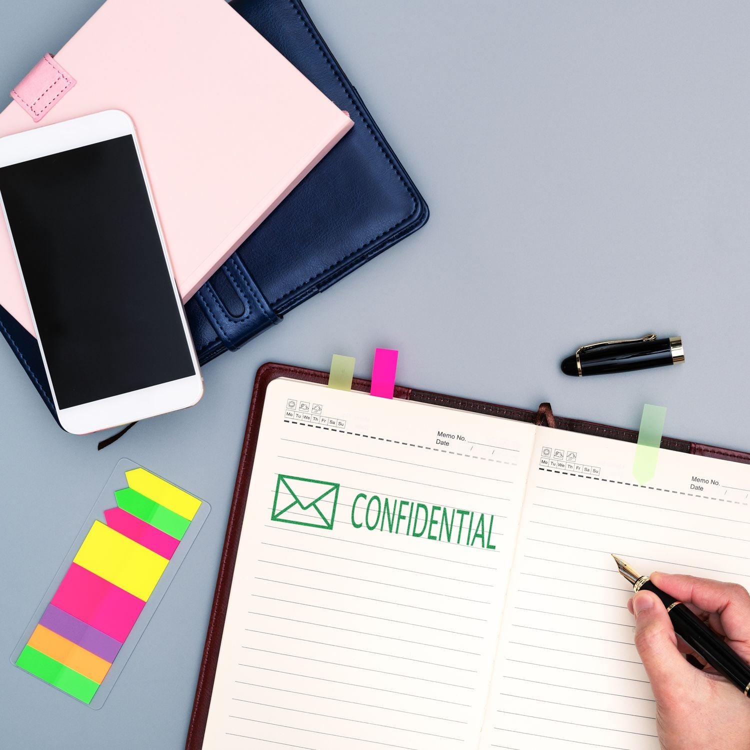 A desk with a notebook stamped CONFIDENTIAL using the Large Self Inking Confidential with Envelope Stamp, alongside a smartphone, colorful sticky notes, and a pen.