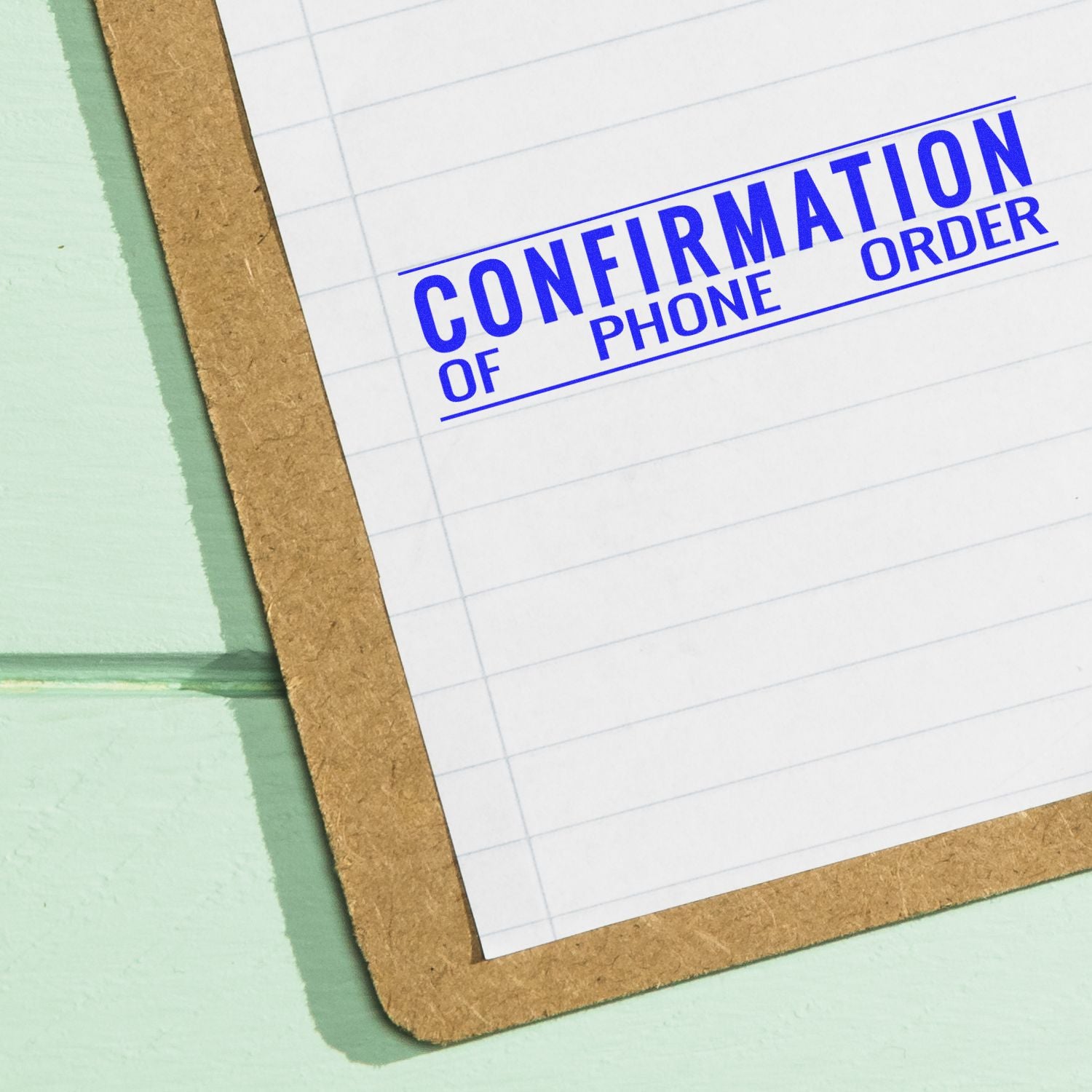 Slim Pre-Inked Confirmation of Phone Order Stamp in blue ink on a lined paper clipped to a brown clipboard on a green surface.