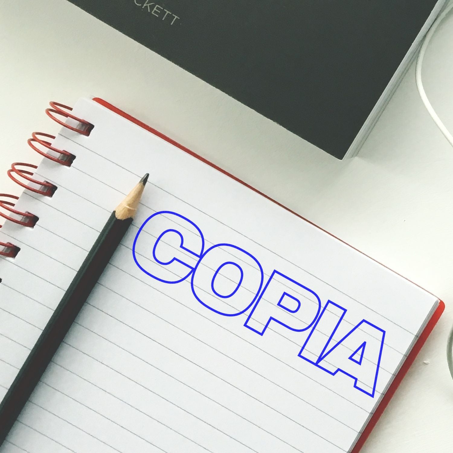 A notebook with a pencil and a bold blue Copia stamped on a page using the Copia Rubber Stamp, placed on a white desk.