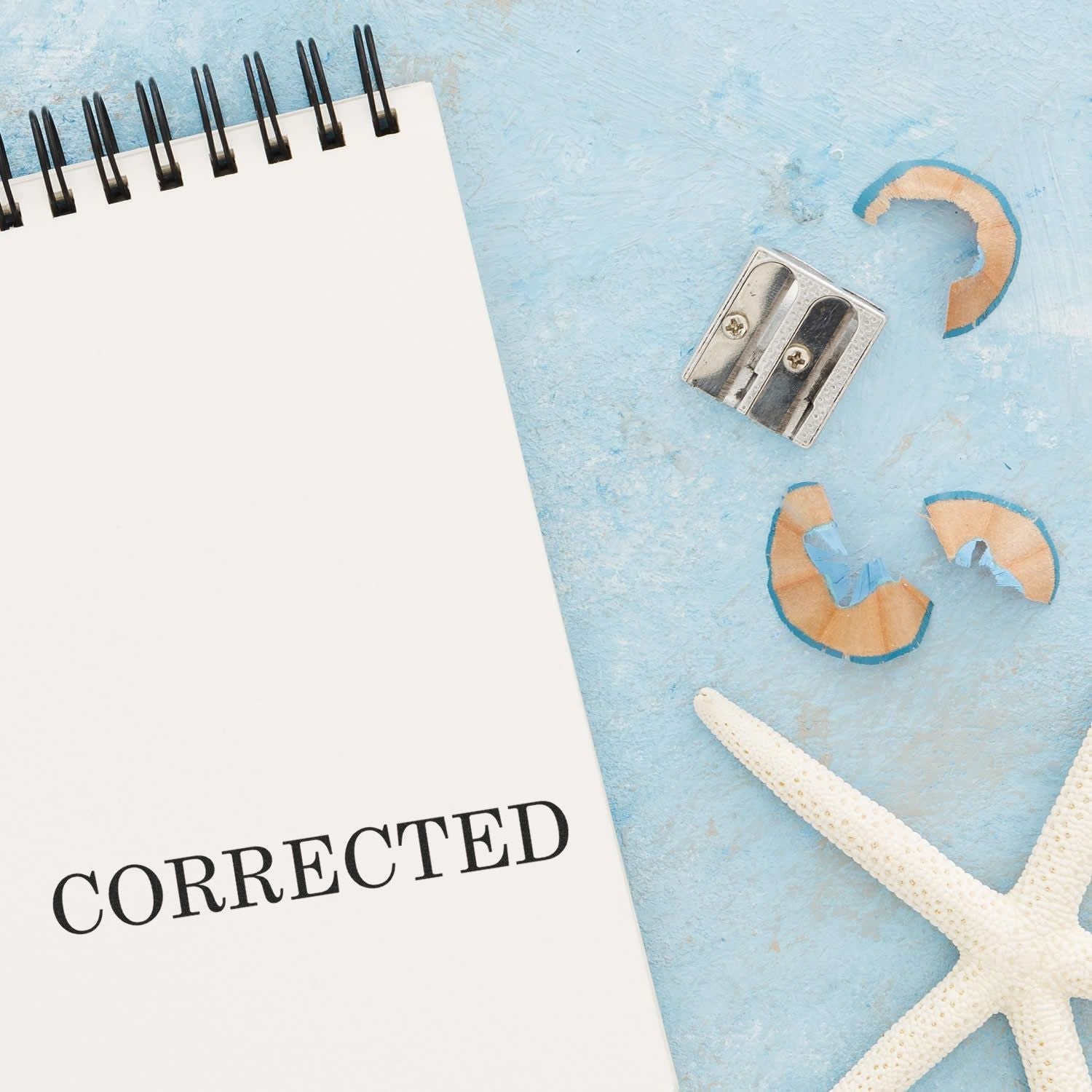 Self Inking Corrected Stamp marking CORRECTED on a notepad, with a pencil sharpener, shavings, and a starfish on a blue surface.