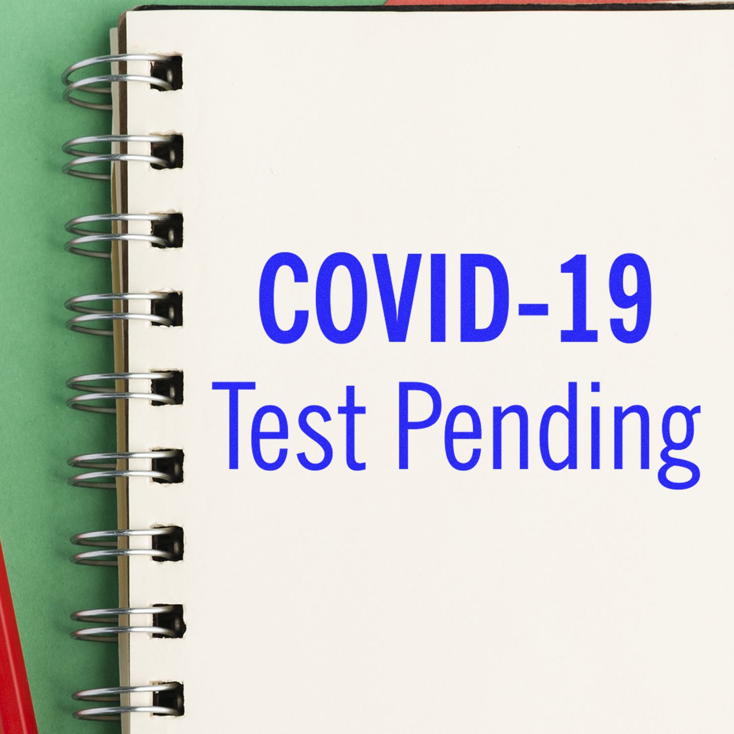 Large Covid-19 Test Pending Rubber Stamp imprint on a spiral notebook, with a red pen beside it on a green surface.