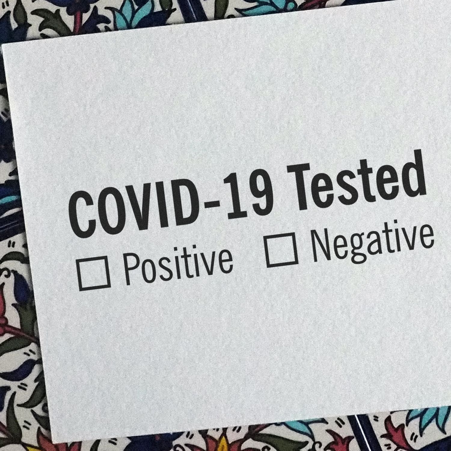 Slim Pre-Inked Covid-19 Tested Stamp on a paper with checkboxes for Positive and Negative results, placed on a floral background.