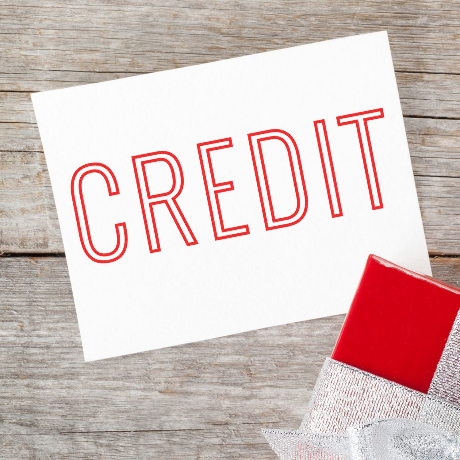 Large Self Inking Credit Outline Stamp used on a white card with CREDIT in red letters, placed on a wooden surface next to a red gift box.