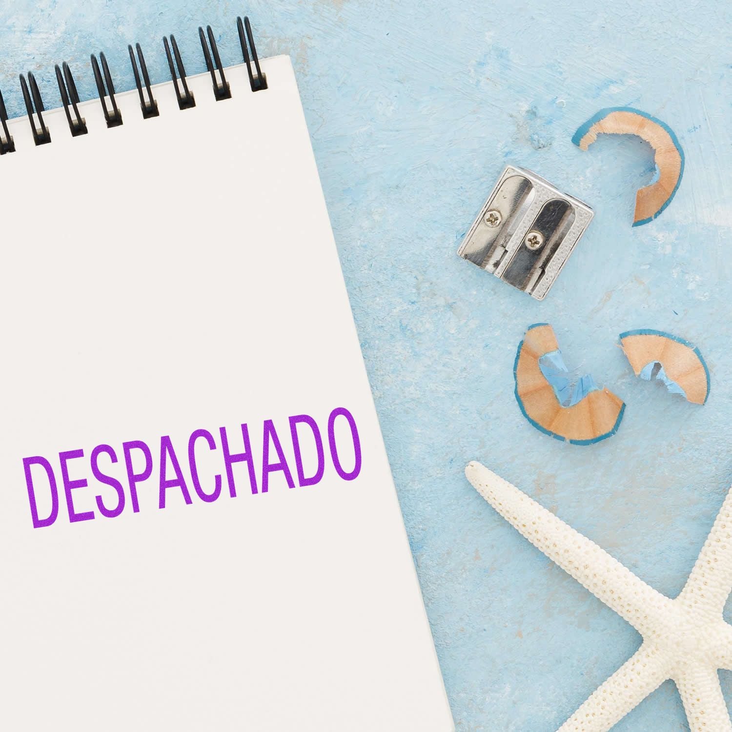 A notebook stamped with DESPACHADO in purple ink using the Large Despachado Rubber Stamp, next to a pencil sharpener and starfish on a blue surface.