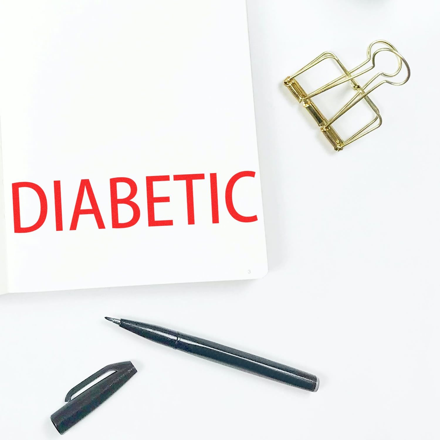 A notebook stamped with DIABETIC using the Diabetic Rubber Stamp, alongside a black pen, pen cap, and a gold binder clip on a white surface.