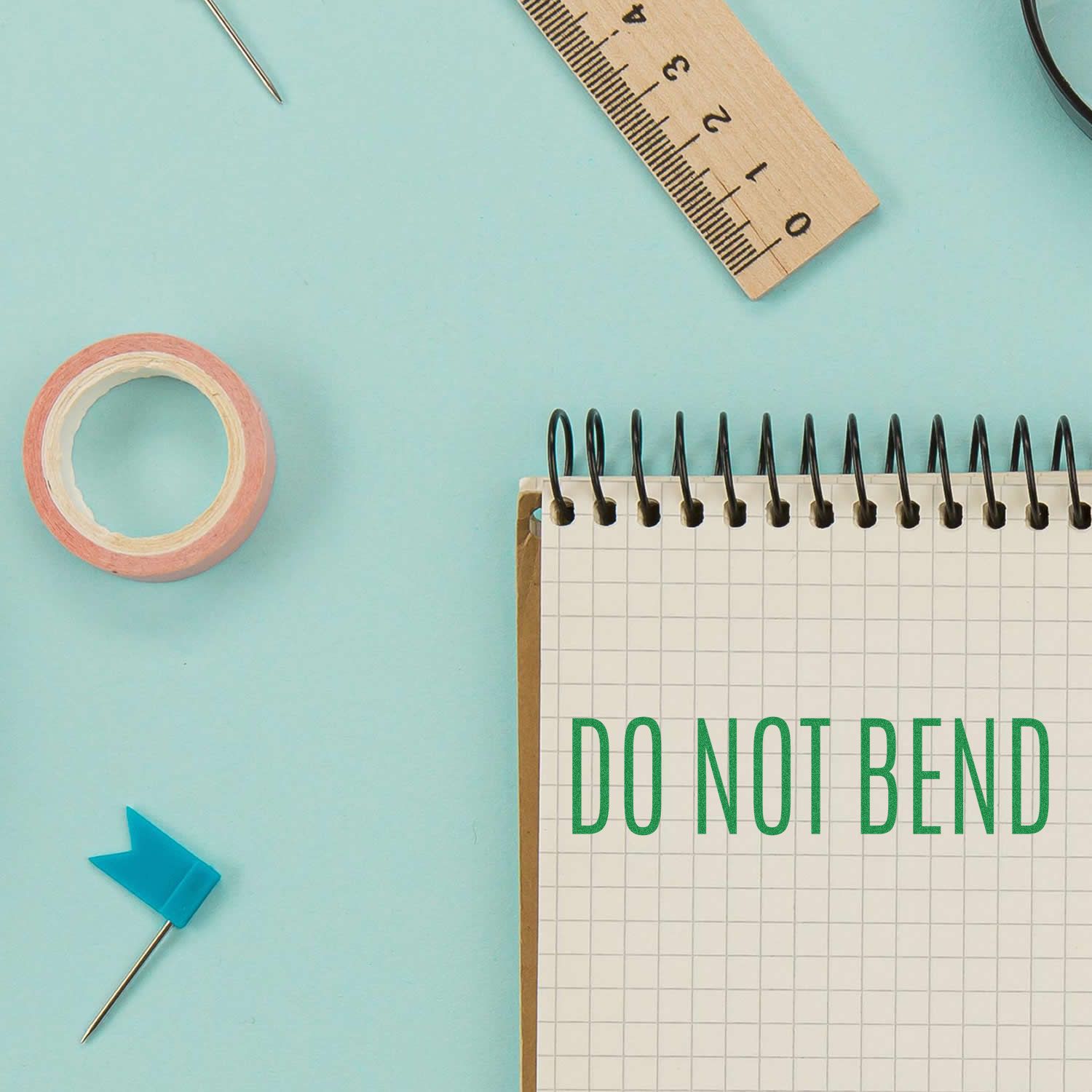 A Do Not Bend Rubber Stamp is shown in use on a spiral notebook, surrounded by a ruler, tape, and a blue flag pin on a light blue background.