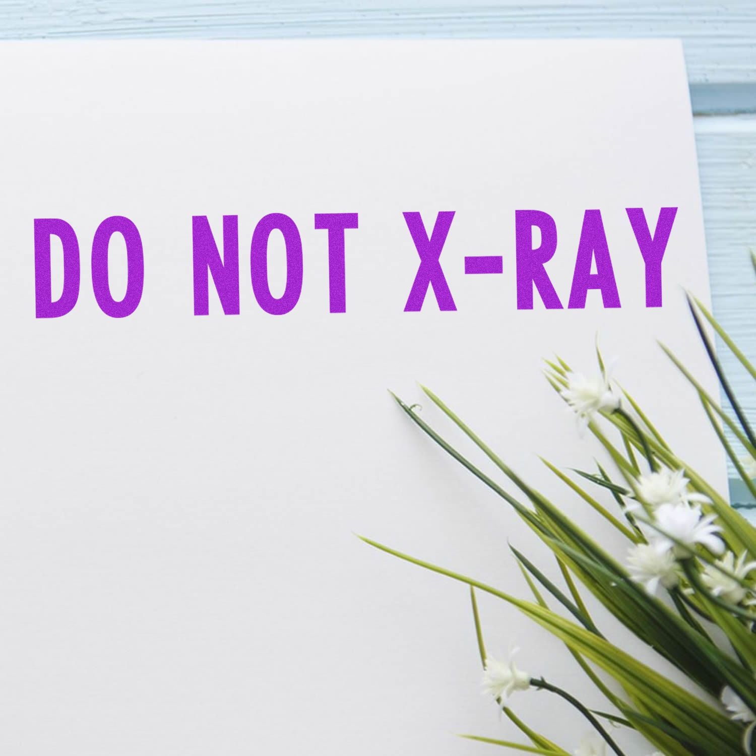 Slim Pre-Inked Do Not X-Ray Stamp in purple ink on white paper, next to green and white flowers on a light blue wooden surface.