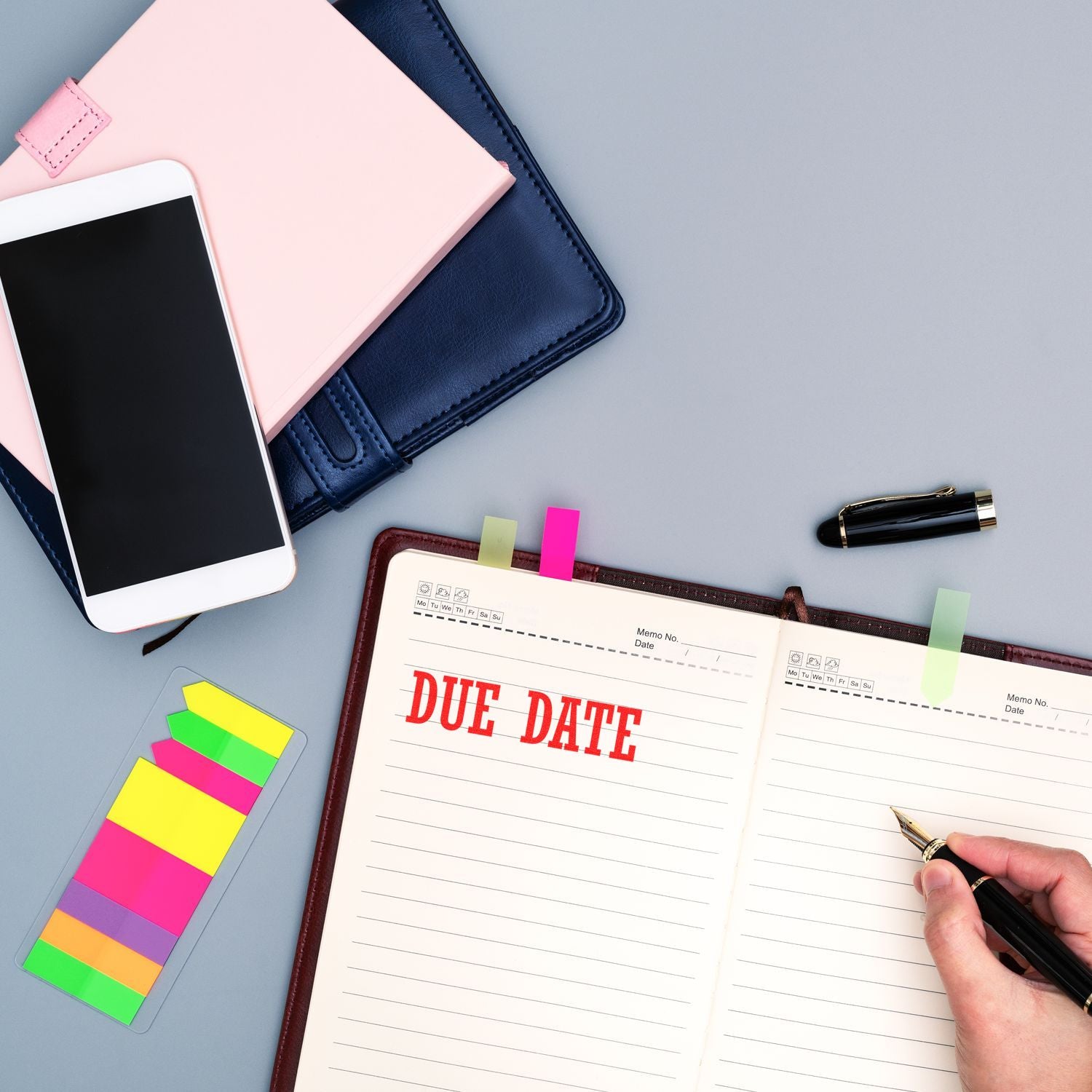 Self Inking Due Date Stamp marking a planner with DUE DATE in red ink, surrounded by notebooks, a phone, sticky notes, and a pen.