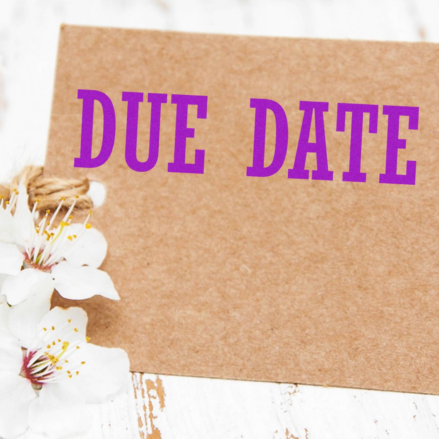 Large Pre-Inked Due Date Stamp in use on a brown card with purple text DUE DATE, next to white flowers on a white wooden surface.