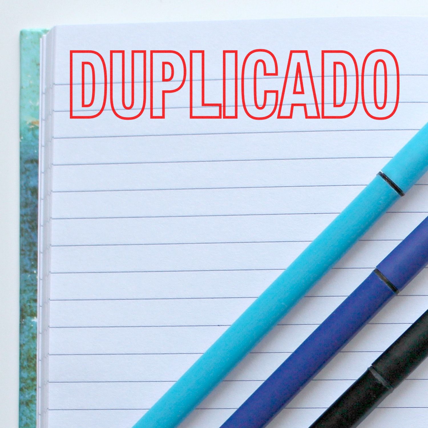 Large Pre-Inked Duplicado Stamp in red ink on a lined notebook page with three pens placed diagonally across the page.