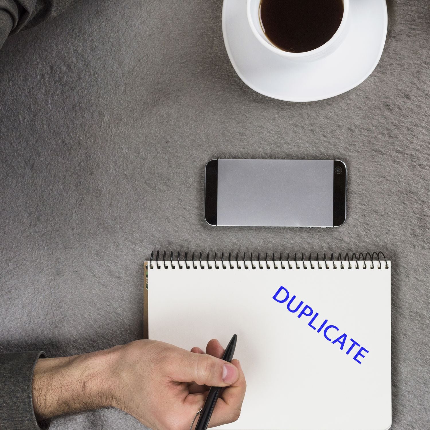 Hand holding pen near notebook stamped with DUPLICATE using Self Inking Duplicate Stamp, with a smartphone and coffee cup on a gray surface.