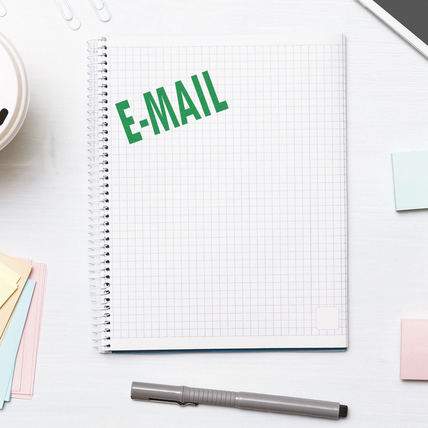 Large Self Inking E Mail Stamp in green ink on a spiral notebook, surrounded by a pen, sticky notes, and a smartphone on a white desk.