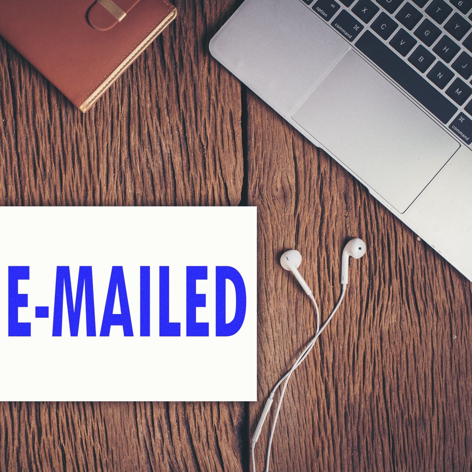 E Mailed Rubber Stamp in blue ink on paper, placed on a wooden desk next to a laptop, earphones, and a brown notebook.