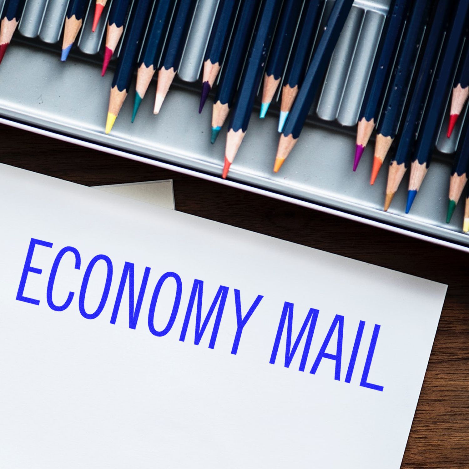 Economy Mail Rubber Stamp in blue ink on a white envelope, with colored pencils in the background."