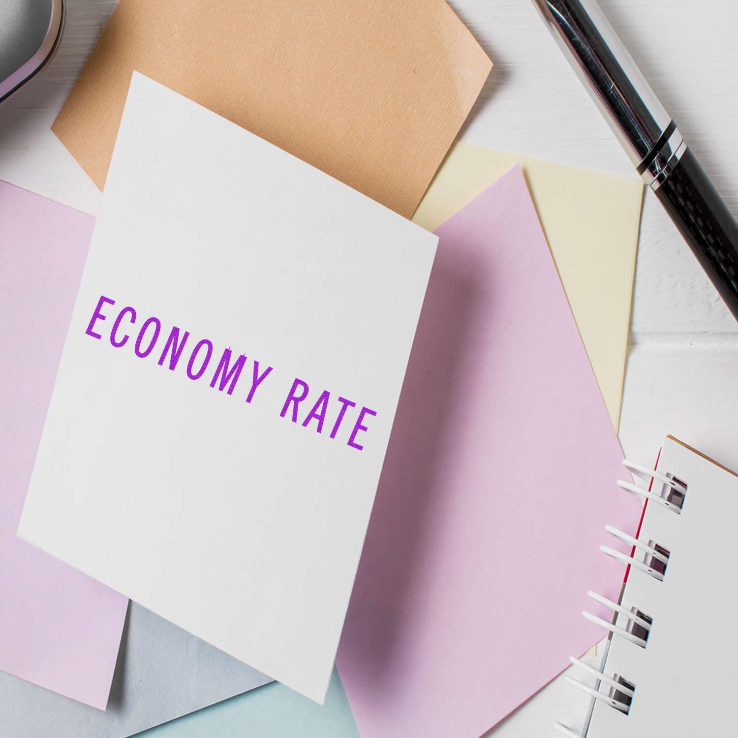 Large Economy Rate Rubber Stamp imprint on a white card surrounded by pastel-colored papers, a pen, and a notebook.