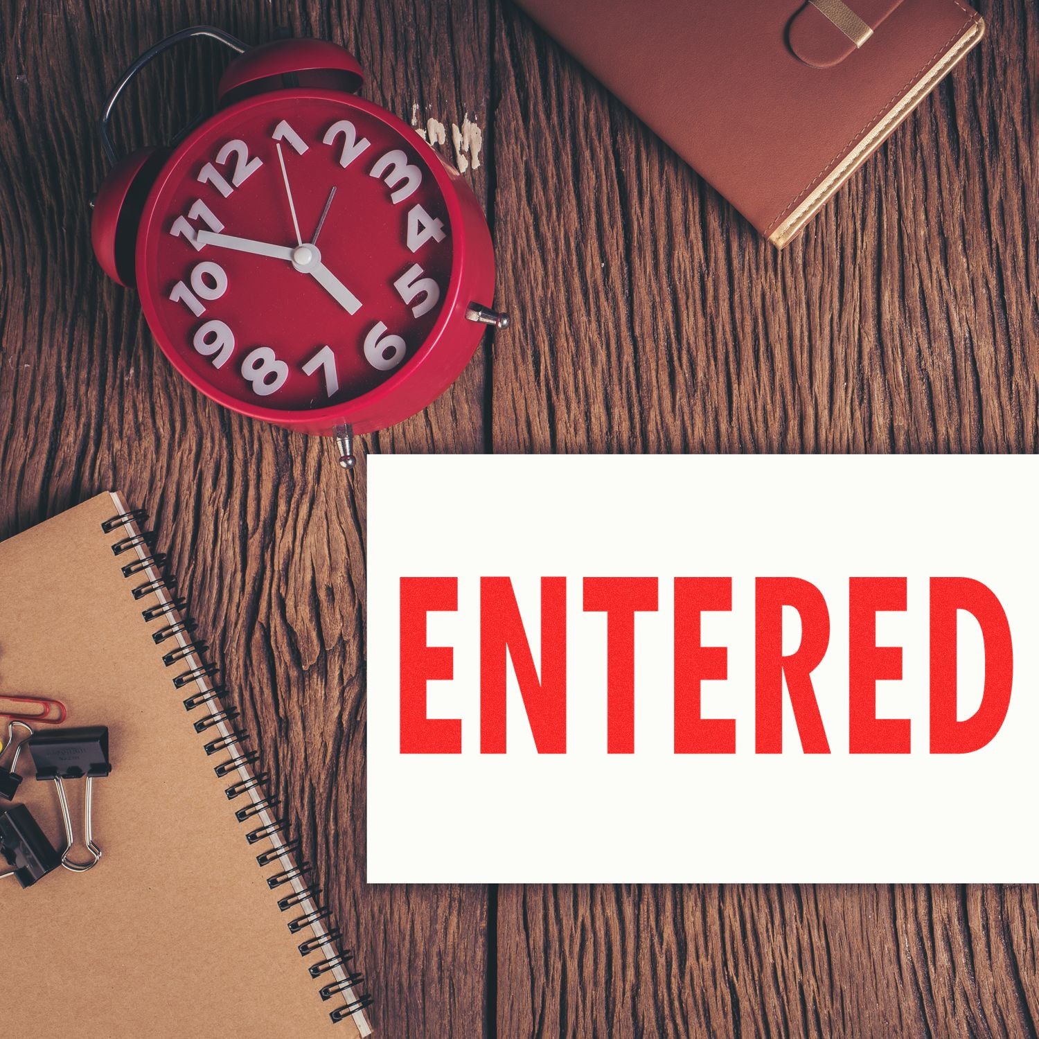 Large Self Inking Entered Stamp in red ink on paper, placed on a wooden desk with a red clock, notebook, and office supplies.