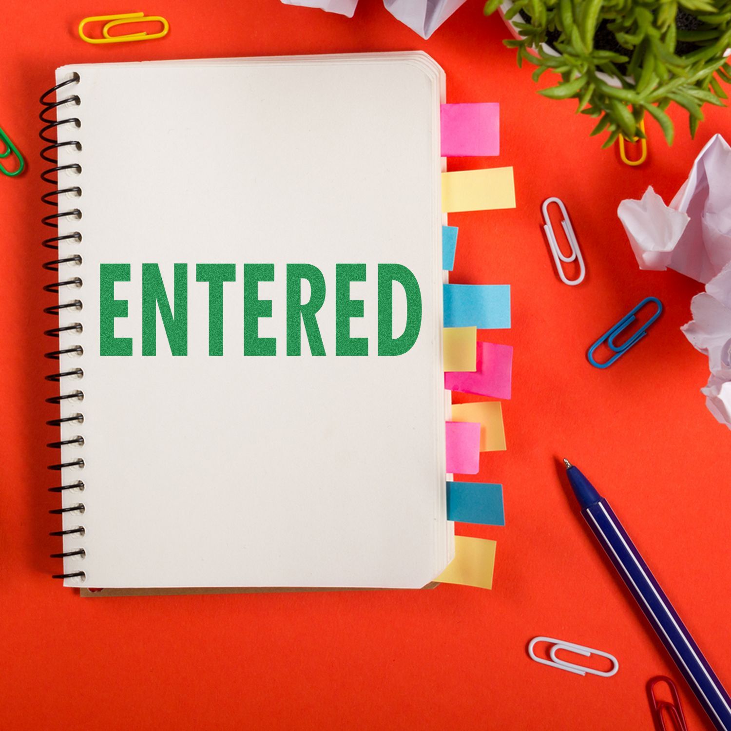 Notebook stamped with ENTERED using the Large Entered Rubber Stamp, surrounded by colorful paper clips, sticky notes, and a pen on a red background.