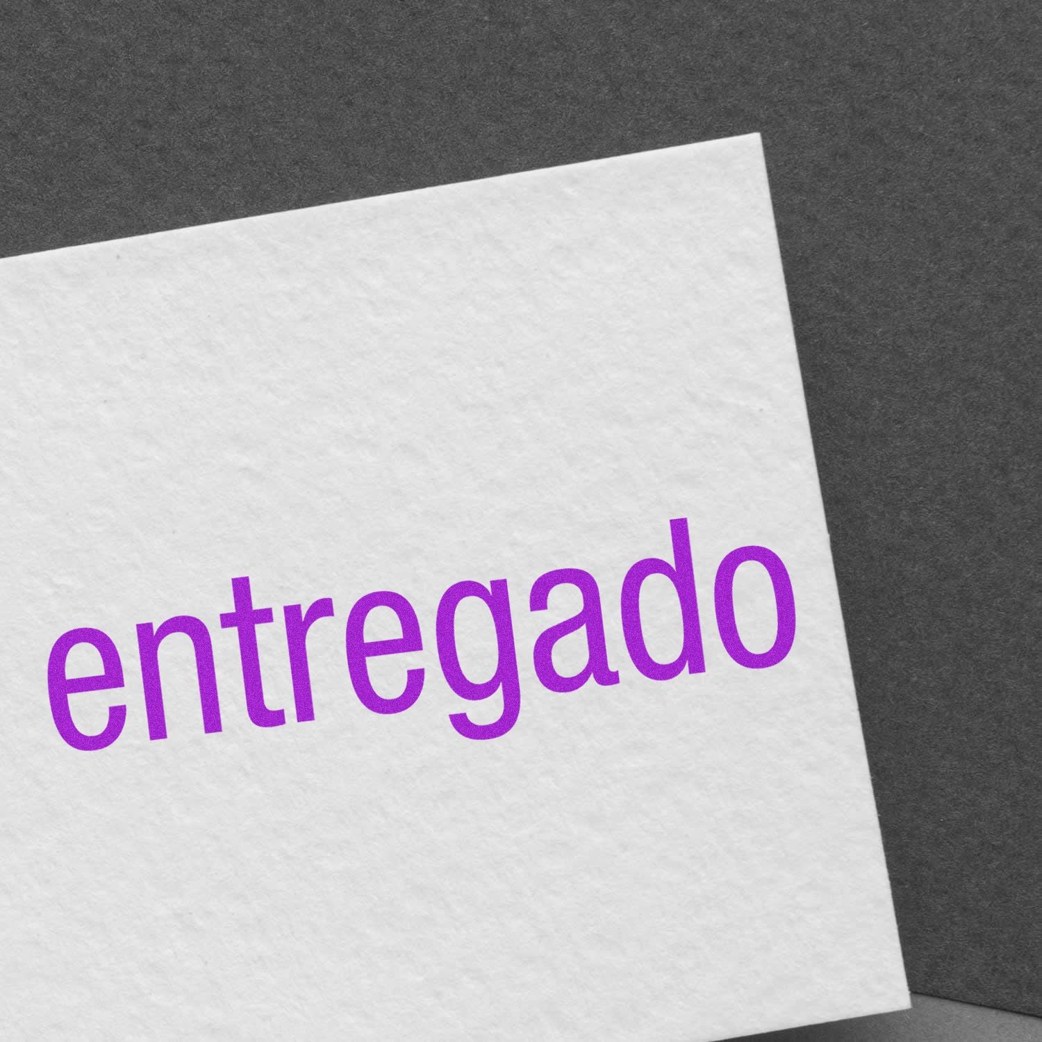 A white card stamped with entregado in purple ink using the Large Entregado Rubber Stamp, placed on a dark gray surface.