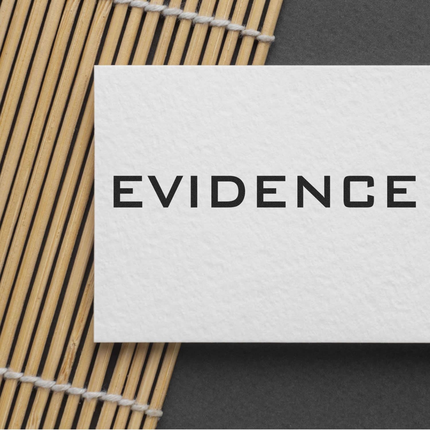 White card stamped with EVIDENCE using the Large Evidence Rubber Stamp, placed on a bamboo mat and dark surface.