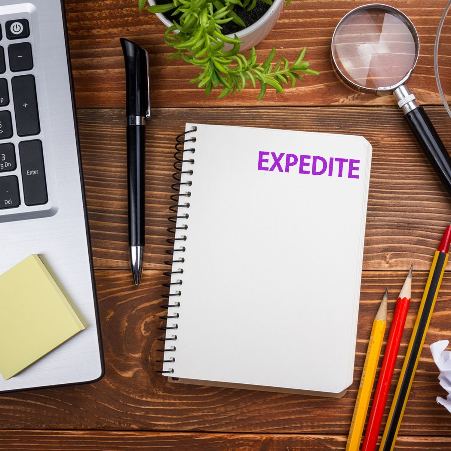 A Self Inking Expedite Stamp is used on a notebook labeled EXPEDITE on a wooden desk with a laptop, pen, magnifying glass, and pencils.