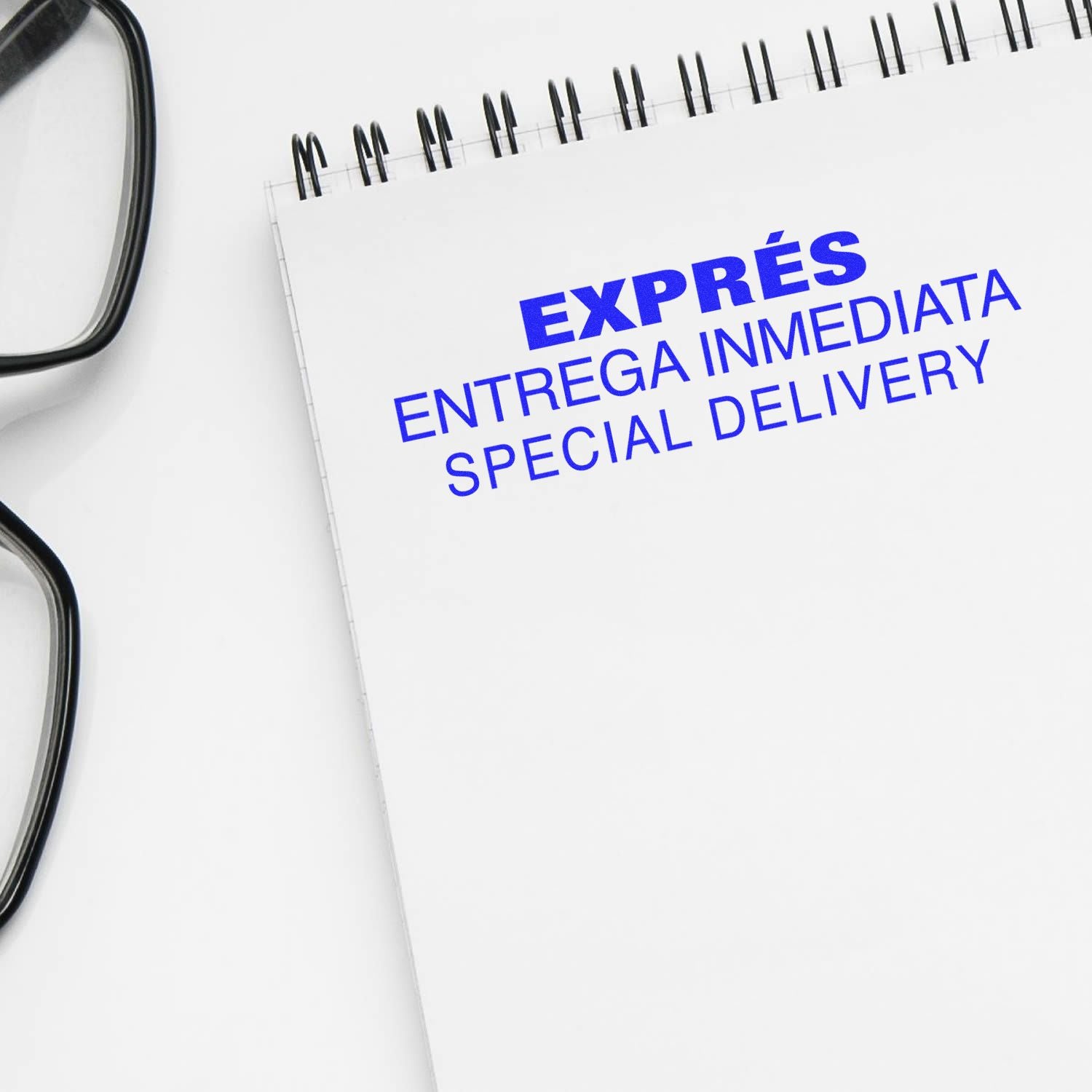 Large Expres Entrega Inmediata Rubber Stamp in blue ink on a white notepad, next to a pair of black glasses.