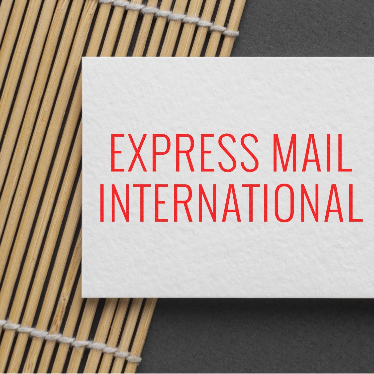 Slim Pre-Inked Express Mail International Stamp in red ink on a white card, placed on a bamboo mat and dark background.