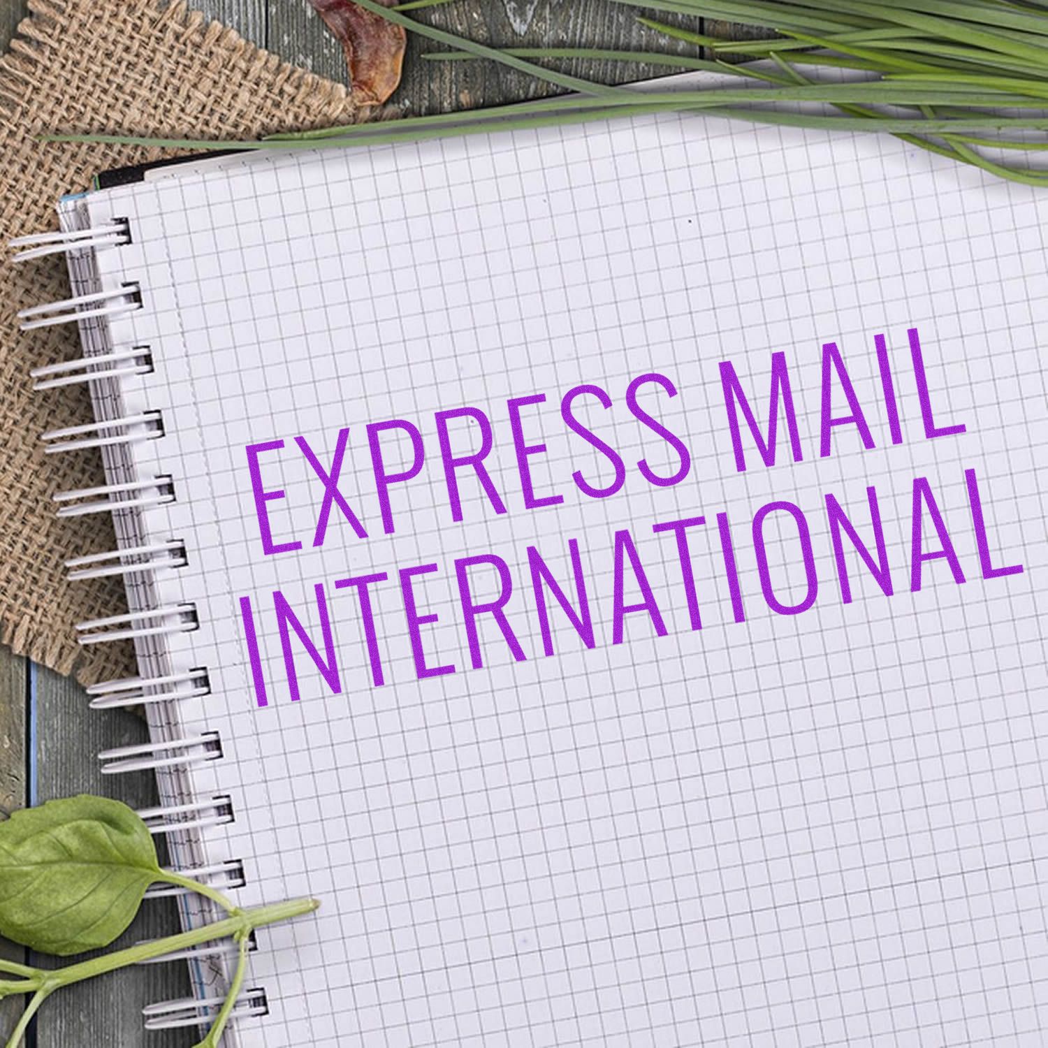 A notebook with Express Mail International stamped in purple using the Express Mail International Rubber Stamp, surrounded by leaves and burlap.