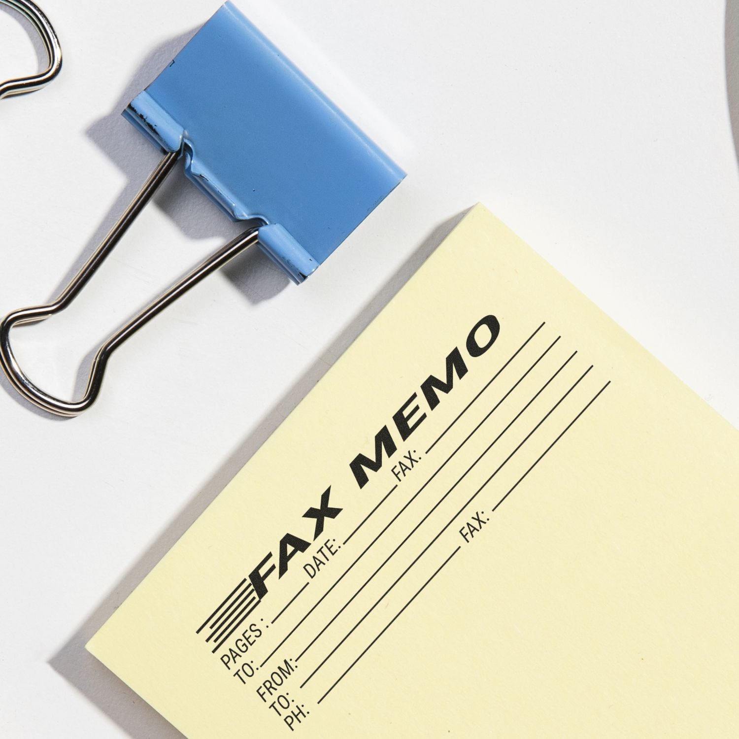 Large Pre-Inked Fax Memo Stamp in use on a yellow memo sheet, accompanied by a blue binder clip on a white surface.