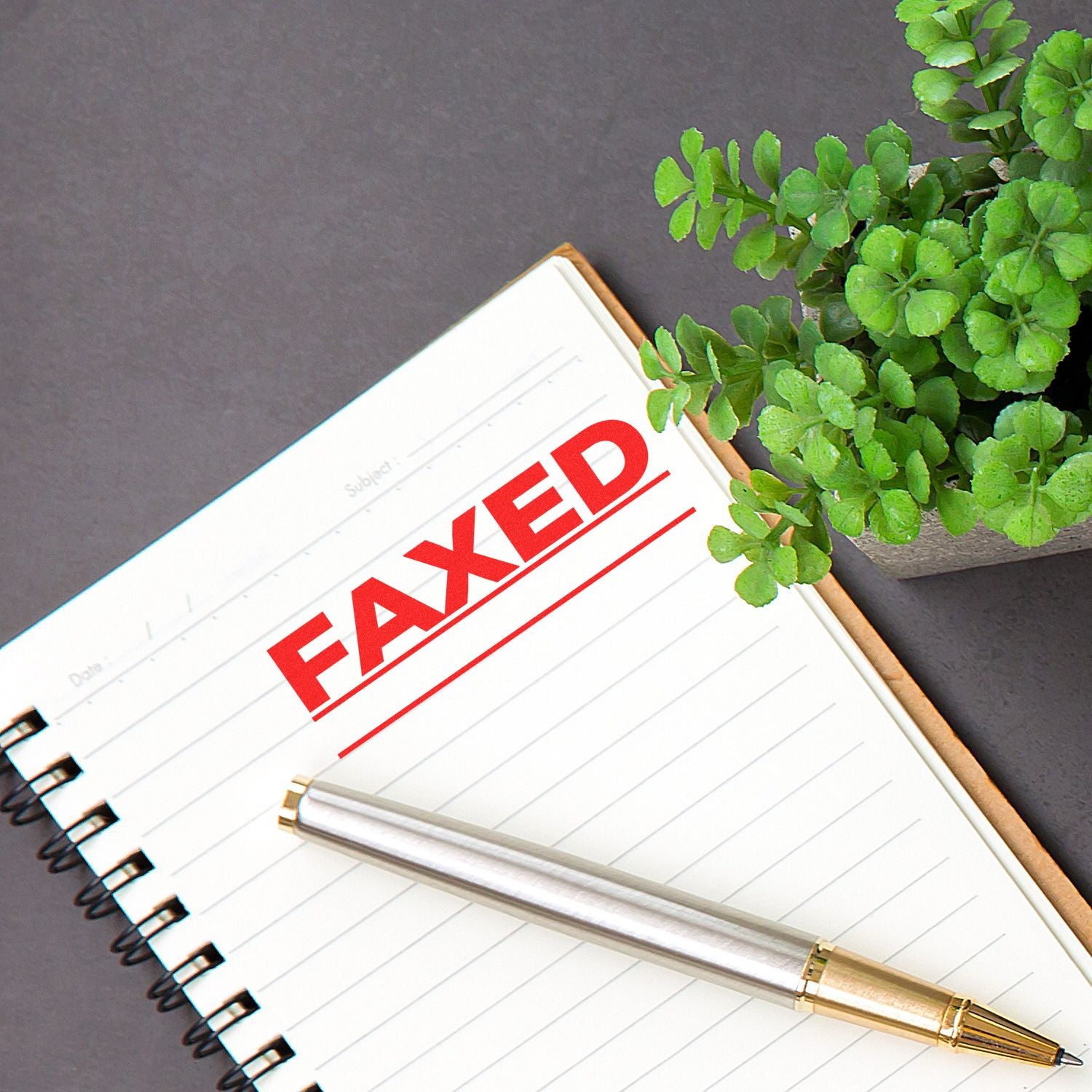 Large Pre-Inked Faxed with Line Stamp in red on a notebook page, next to a silver pen and a small green plant on a dark surface.