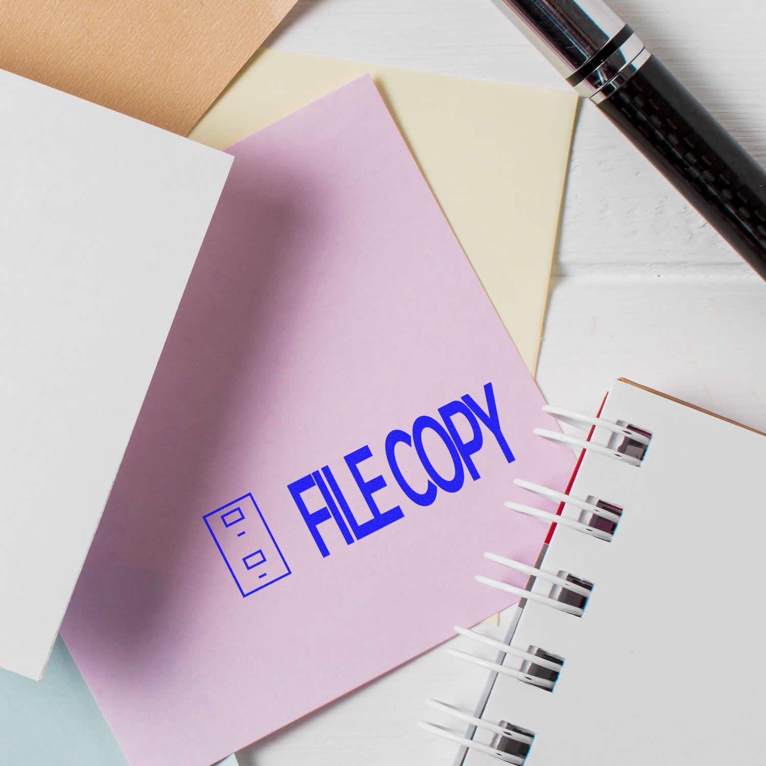 File Copy with Drawer Rubber Stamp used on a pink paper, surrounded by other papers, a pen, and a notebook on a white surface.