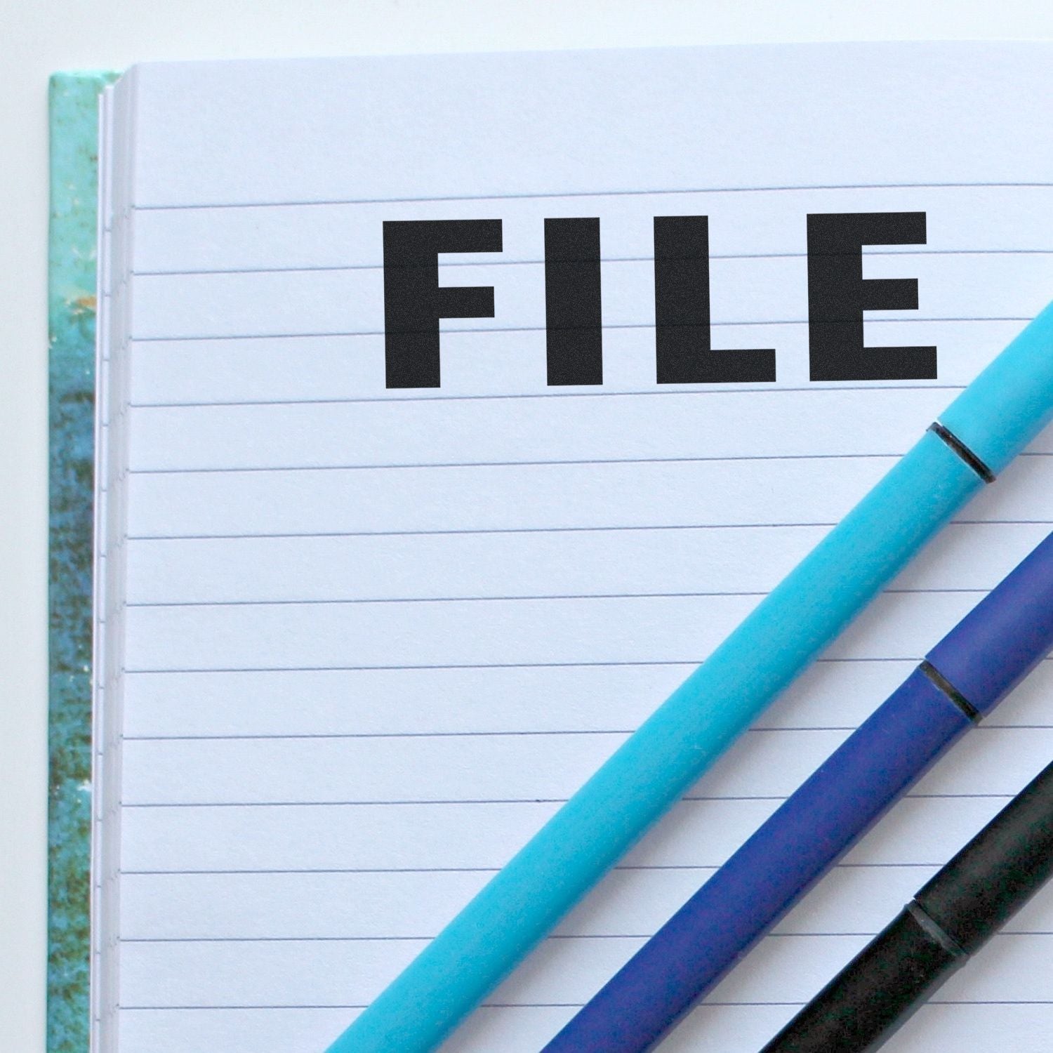 Self Inking File Stamp marking the word FILE on a lined notebook page, with two pens placed diagonally across the page.