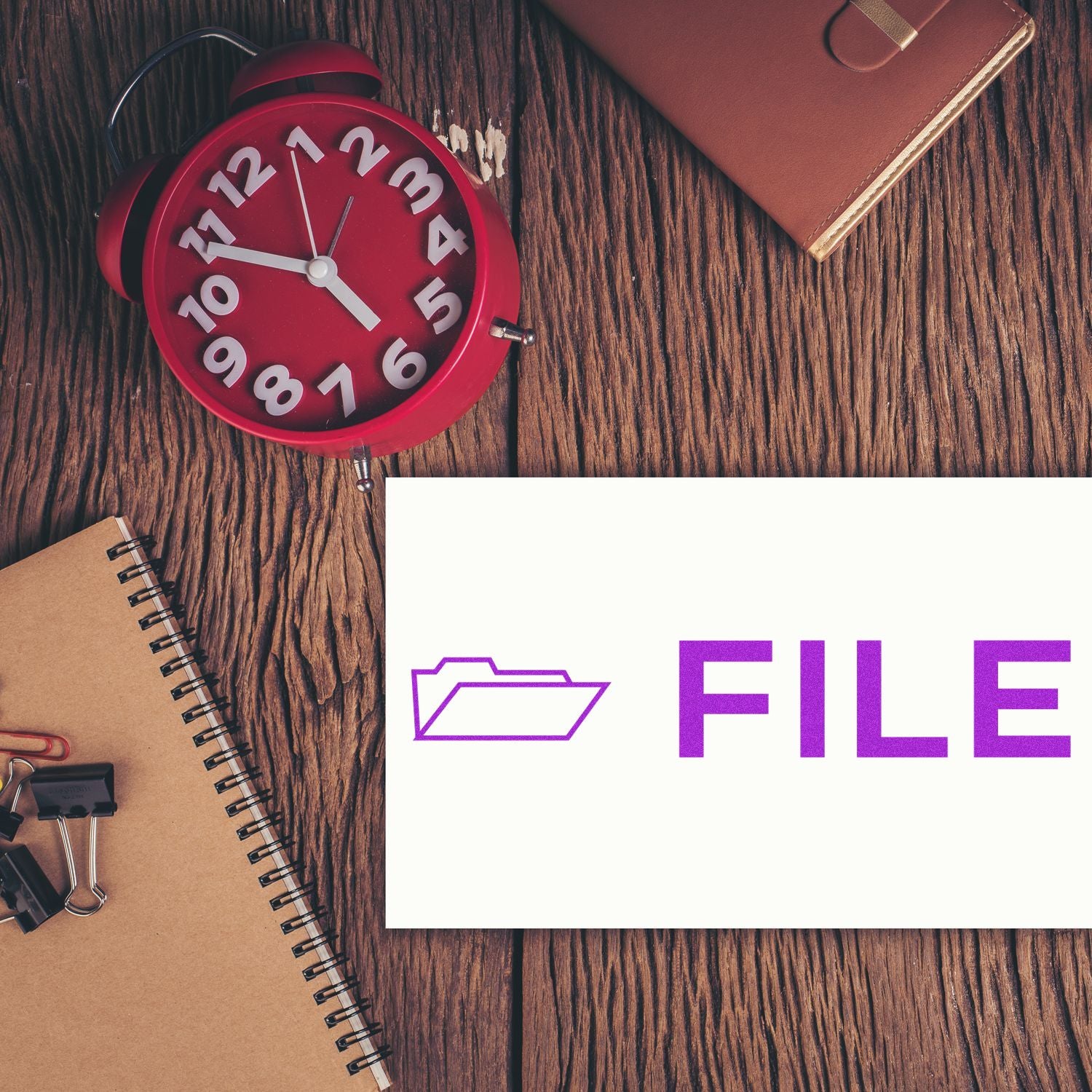 Large File with Envelope Rubber Stamp on a white card, placed on a wooden desk with a red clock, notebook, and office supplies.