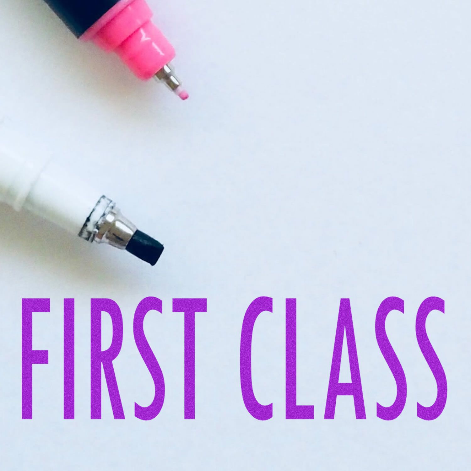 First Class Mailing Rubber Stamp in use, with FIRST CLASS text stamped in purple ink on white paper, next to two marker pens.