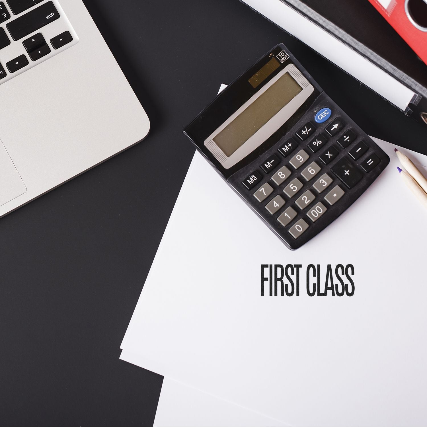 A desk with a laptop, calculator, and papers stamped with First Class using the First Class Rubber Stamp.