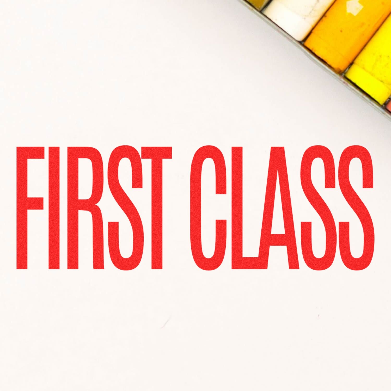 Red 'FIRST CLASS' text stamped on white paper using a Slim Pre-Inked First Class Stamp, with a colorful object in the corner.